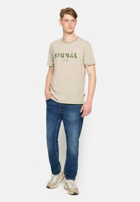 Camiseta de rayas beige con el texto verde "SIGNAL", combinada con jeans azules y zapatillas claras, de pie de manera casual con una mano en el bolsillo.