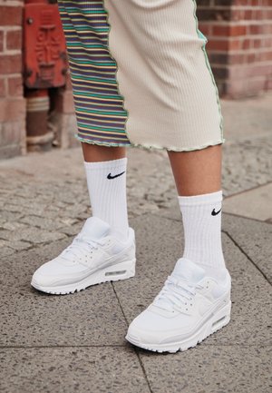 Nogi w białych butach sportowych Nike i białych skarpetkach Nike stojące na brukowanym chodniku obok ceglanej ściany.