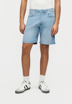 Mand iført lyseblå denimshorts, hvide sokker og hvide sneakers med sorte striber, foran en ensfarvet lys baggrund.
