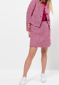 Veste en tweed rose avec encolure ronde et jupe assortie, présentant un motif texturé et des boutons. Associée à des baskets blanches.
