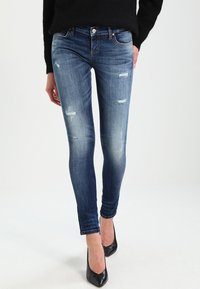 Jeans ajustados en denim azul oscuro, con un ligero desgastado y parches rasgados. Combinados con tacones altos negros para crear contraste.