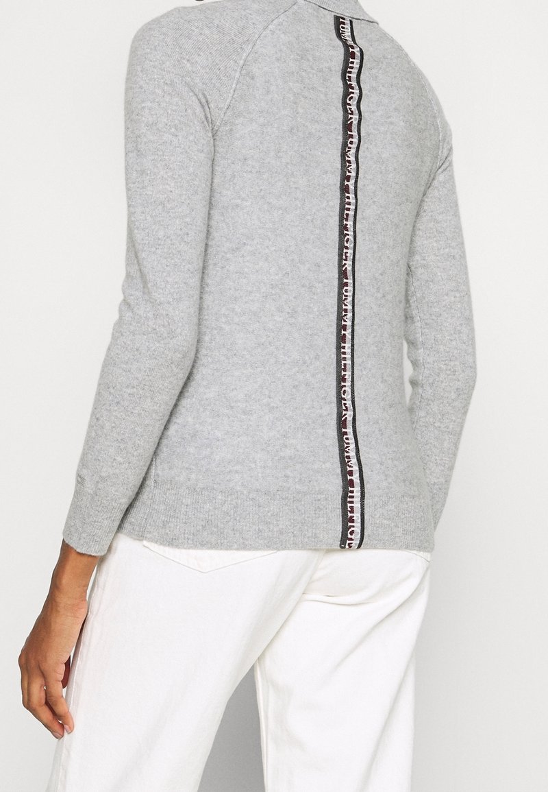 Personne portant un pull à manches longues gris clair avec une bande verticale noire et rouge de marque dans le dos, associée à un pantalon blanc.