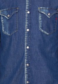 Camicia in denim di colore blu scuro con una texture sbiadita, dotata di bottoni a pressione argentati e due tasche sul petto con alette appuntite.