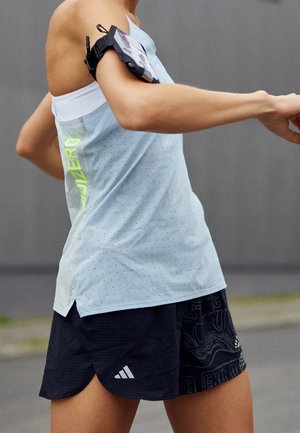 Biegacz ubrany w jasnoniebieską koszulkę bez rękawów i czarne spodenki Adidas, z czarną opaską na górnej części ramienia trzymającą smartfon.