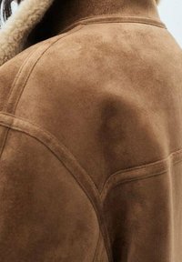 Veste en daim marron avec une texture douce et des détails de couture visibles. Elle possède un large col doublé d'un matériau plus clair et duveteux.