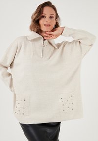 Pullover beige clair avec un col côtelé et une fermeture éclair mi-longue. Présente deux poches avant ornées de petites clous métalliques.