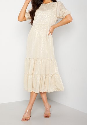 Kobieta w kremowej, teksturowanej sukience midi z bufkami oraz falbanami na dole, w połączeniu z przezroczystymi sandałami na wysokim obcasie ze paskiem.