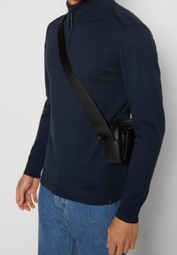 Uomo che indossa un maglione blu navy con zip, una borsa a tracolla nera e jeans blu, su sfondo uniforme.