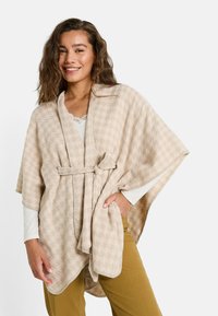 Poncho beige à motifs en tissu doux, doté d'un lien à la taille et d'un col châle, porté sur une chemise blanche à manches longues.