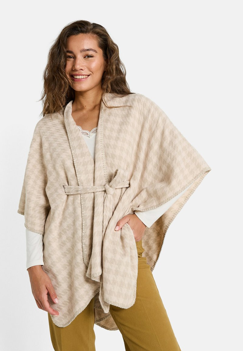 Poncho beige à motifs en tissu doux, doté d'un lien à la taille et d'un col châle, porté sur une chemise blanche à manches longues.
