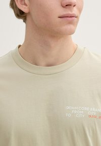 Lichtbeige t-shirt met een ronde hals, voorzien van een minimalistisch tekstopdruk in het wit en rood op de borst, gemaakt van zachte stof.