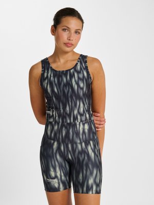 Vrouw die een mouwloos, zwart-wit patroon atletisch crop top en bijpassende shorts met hoge taille draagt, staand met één arm achter haar rug.