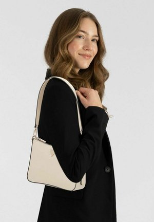 Joven con cabello ondulado que lleva un blazer negro y un pequeño bolso beige de hombro, sonriendo ligeramente y de perfil.