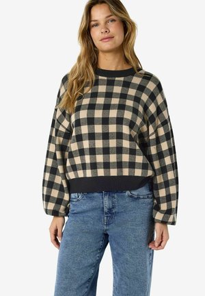 Femme portant un pull beige et noir à carreaux avec des manches bouffantes et un jean bleu, debout devant un fond blanc uni.