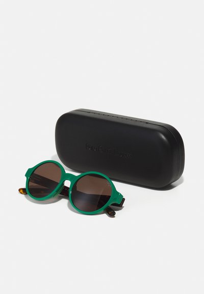 Lunettes de soleil rondes vertes avec des verres marron et une finition brillante. Accompagnées d'un étui rigide noir mat avec un marquage en relief.