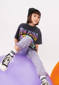 Črna kapa, grafična siva majica Pink Floyd, izbljena modra jeans hlače, črne superge z belimi podplati, sedi na veliki vijolični žogi.