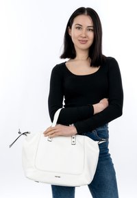 Ung kvinde med glat mørkt hår iført sort top og blå jeans, der holder en stor hvid håndtaske med dobbelte håndtag.