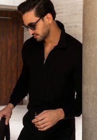 Sudadera negra con cremallera, con un patrón texturizado, mangas largas y cuello, combinada con pantalones negros. Los accesorios incluyen gafas de sol.