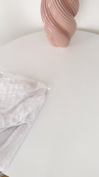 Hvite blondetrim undertøy pakket i klar plast, plassert på et rundt hvitt bord, ved siden av en rosa, svunget keramikkvase.