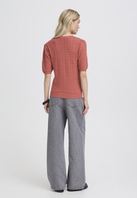 Frau steht mit dem Rücken zur Kamera, trägt ein strukturiertes, korallenfarbenes Kurzarmtop und weite graue Jeans vor einem schlichten Hintergrund.