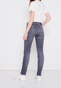 Mujer que lleva jeans ajustados grises, zapatillas blancas y una camiseta blanca, de pie con una mano en la cadera contra un fondo blanco liso.