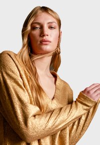 Guld, metallisk stickad tröja med V-ringning, långa ärmar och texturerad finish. Håret stylat med mjuka vågor, minimal smyckespynt.