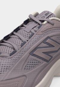 Κοντινό πλάνο ενός γκρι παπουτσιού New Balance με ύφασμα από δίχτυ, επενδεδυμένο γιακά και λεπτομερή κορδόνια σε λευκή σόλα.