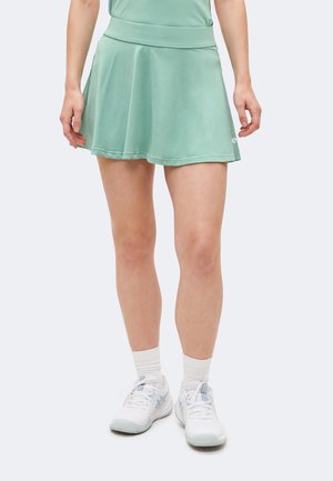 Žena v svetlozelenej športovej sukni, bielych ponožkách a bielych športových topánkach stojí na jednofarebnom svetlom pozadí.