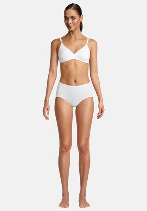 Woman standing barefoot in white wireless bra and high-waisted briefs against plain white background, looking slightly right.