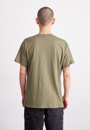 Homme à la tête rasée portant un t-shirt vert olive à manches courtes et un pantalon cargo foncé, vu de dos sur un fond uni.