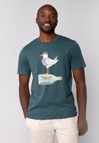 T-shirt en coton turquoise à manches courtes pour homme, présentant un graphique d'un oiseau blanc en chemise rayée se tenant sur une bouteille avec un bateau en papier à l'intérieur.