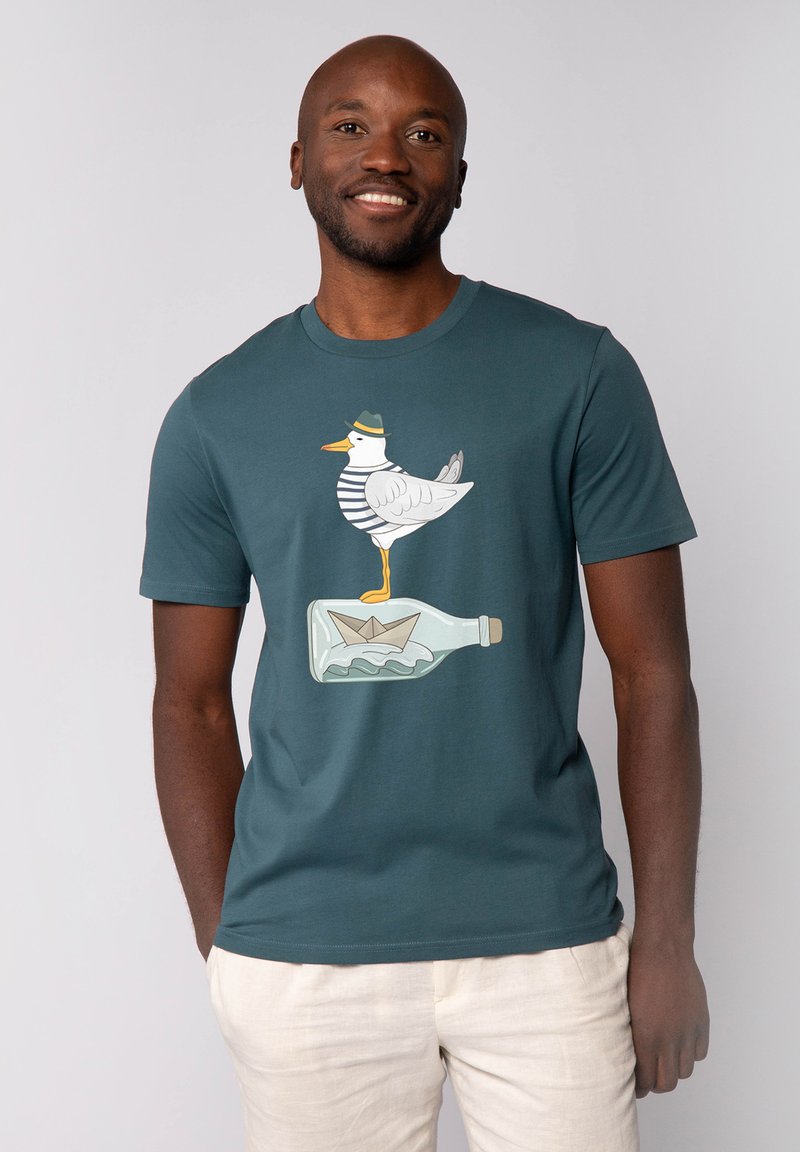 T-shirt en coton turquoise à manches courtes pour homme, présentant un graphique d'un oiseau blanc en chemise rayée se tenant sur une bouteille avec un bateau en papier à l'intérieur.