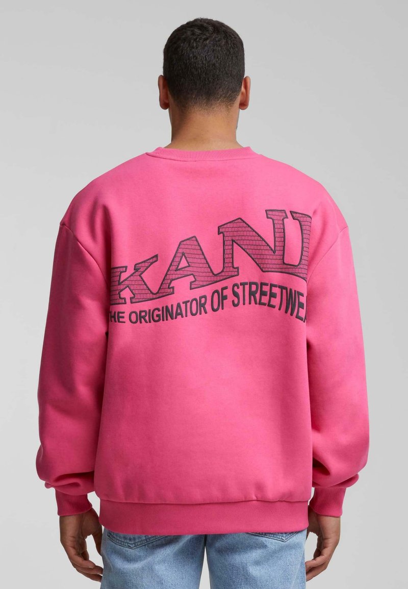 Rosa sweatshirt med ribbade ärmar och nederkant, med stor svart text på baksidan: "KANDI ORIGINALAREN AV STREETWEAR."