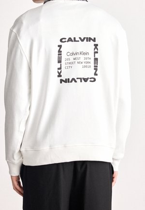 Sweat-shirt blanc avec inscription noire Calvin Klein formant un carré autour d'une adresse à New York dans le dos, porté par une personne debout.
