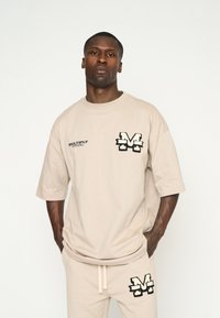 Camiseta oversized beige de manga corta con logo negro bordado en el pecho. Combinada con joggers a juego con un detalle de logo similar.