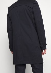 Mörkblå lång kappa med rak skärning, knäppta ärmslut och en mjuk yta. Bakdelen har en ren design med minimala sömmar och detaljer.