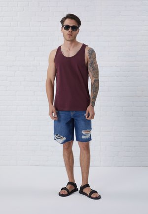 Burgunderfarget topp, revne denimshorts og svarte sandaler. Har en avslappet passform og en tatovering på høyre arm. Hvit teglsteinsbakgrunn.