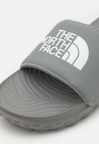 Grå slide-sandal med en texturerad gummisula, som har ett brett rem med präglingen "THE NORTH FACE" i vitt.