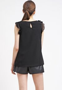 Haut noir sans manches avec détails volantés aux épaules, ouverture en trou de serrure dans le dos, et tissu lisse et léger. Porté avec un short noir.