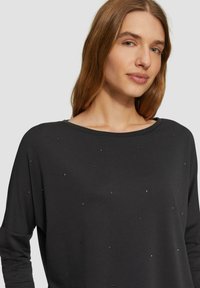 Schwarzes Langarmshirt mit weitem U-Ausschnitt, verziert mit verstreuten metallischen Akzenten. Weicher Stoff mit entspannter Passform und überschnittenen Schultern.