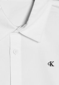 Vit skjorta med klassisk krage, prydd med en liten svart "cK"-logotyp på vänster bröst, tillverkad av mjukt bomullstyg.