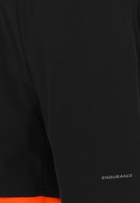 Svarta träningsshorts med en orange detalj längst ner. Har en jämn yta och en reflekterande "ENDURANCE"-logotyp på sidan.