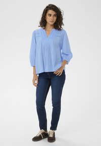 Blouse bleu clair à manches trois-quarts, col en V et détails plis piqués. Associée à un jean en denim foncé et des baskets marron.