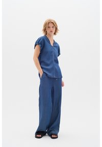 Denimový outfit s vrškem s V-výstřihem a nařasenými rukávy a kalhotami širokého střihu. Tkanina je texturovaná, v tmavě modré barvě s viditelným šitím.