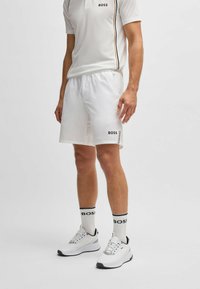 Camisa polo blanca con rayas negras y doradas, pantalones cortos blancos con el logo "BOSS", calcetines en blanco y negro, y zapatillas deportivas blancas.