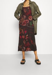 Robe midi noire avec un motif fleuri multicolore, portée sous une veste en cuir olive ajustée. Des sandales à plateforme noires complètent le look.