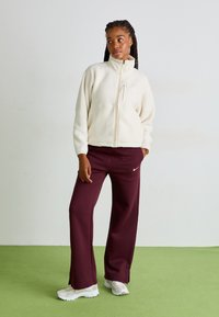 Krämfärgad fleece zip-up jacka med hög krage, tillsammans med vinröda sweatpants med vitt Nike-logotyp och vita sportskor.