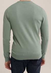 Zelený pletený svetr s dlouhými rukávy, kulatým výstřihem a žebrovanými manžetami a lemem. Nosený přes tmavé džíny, které ukazují hladký, texturovaný povrch.