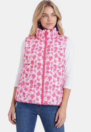 Donna che indossa un gilet imbottito floreale rosa e bianco sopra una camicia bianca a maniche lunghe e jeans blu, guarda di lato con orecchini visibili.