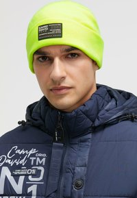 Neon yellow knit beanie with a black label. Model wears a navy puffer jacket featuring logos and accents, showcasing a quilted texture.